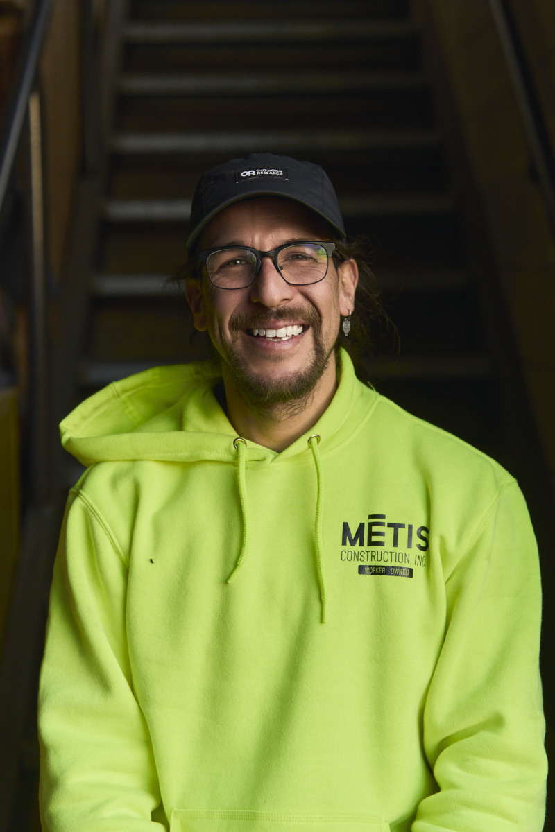 20260120_Metis__DSC1883 Smiling man in a neon green METIS Construction hoodie, glasses, and a dark cap, standing in front of a staircase.