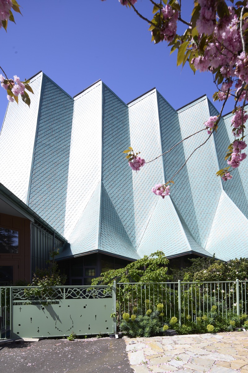 20260425_Metis_StPaul_4221 Modern building with angular light-blue metal roof and a green fence, framed by pink cherry blossoms in bloom against a clear blue sky.