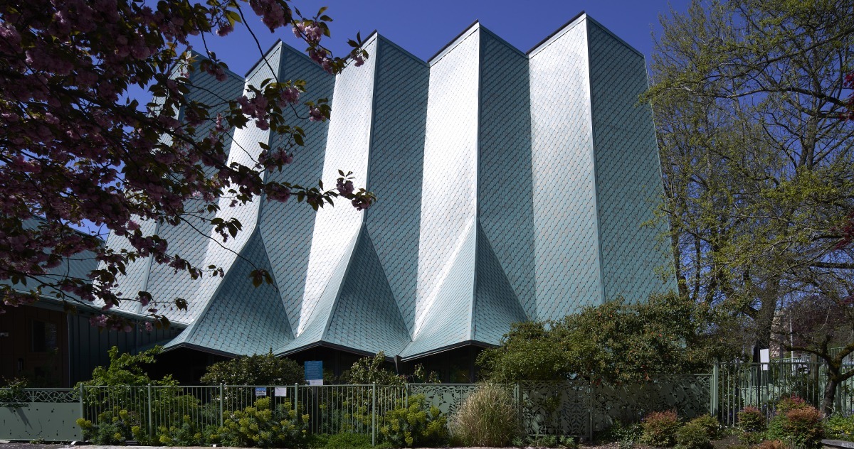 20260425_Metis_StPaul_4222 Angular teal metal roof building with zigzag peaks, surrounded by green shrubs and a fence.