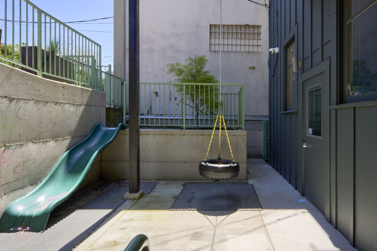 20260425_Metis_StPaul_4237 Tire swing hanging from chains over a padded area in a small playground, with a green slide on the left and a dark blue building on the right.
