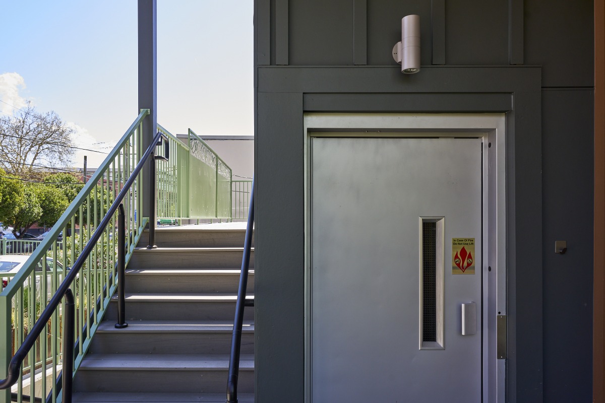 20260425_Metis_StPaul_4242 Exterior metal fire door with a vertical window, beside an outdoor staircase with green railing and black handrails, daylight day.