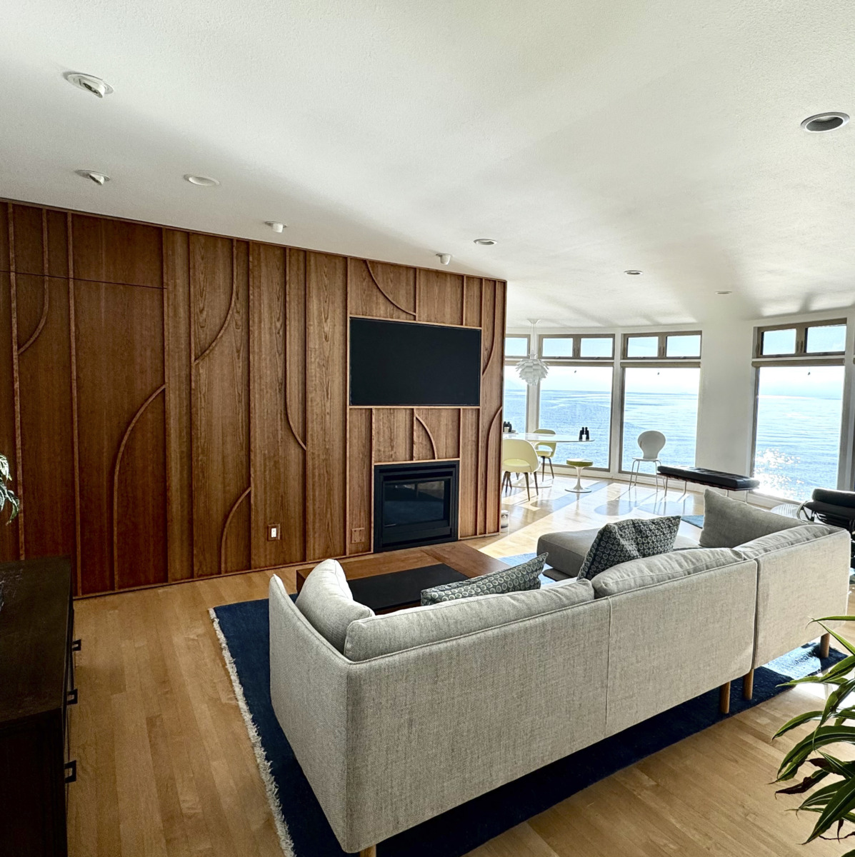 image0 (15)_edited1 Bright oceanfront living room with a wood-paneled accent wall, fireplace, and wall-mounted TV, plus a gray sectional sofa facing large windows.
